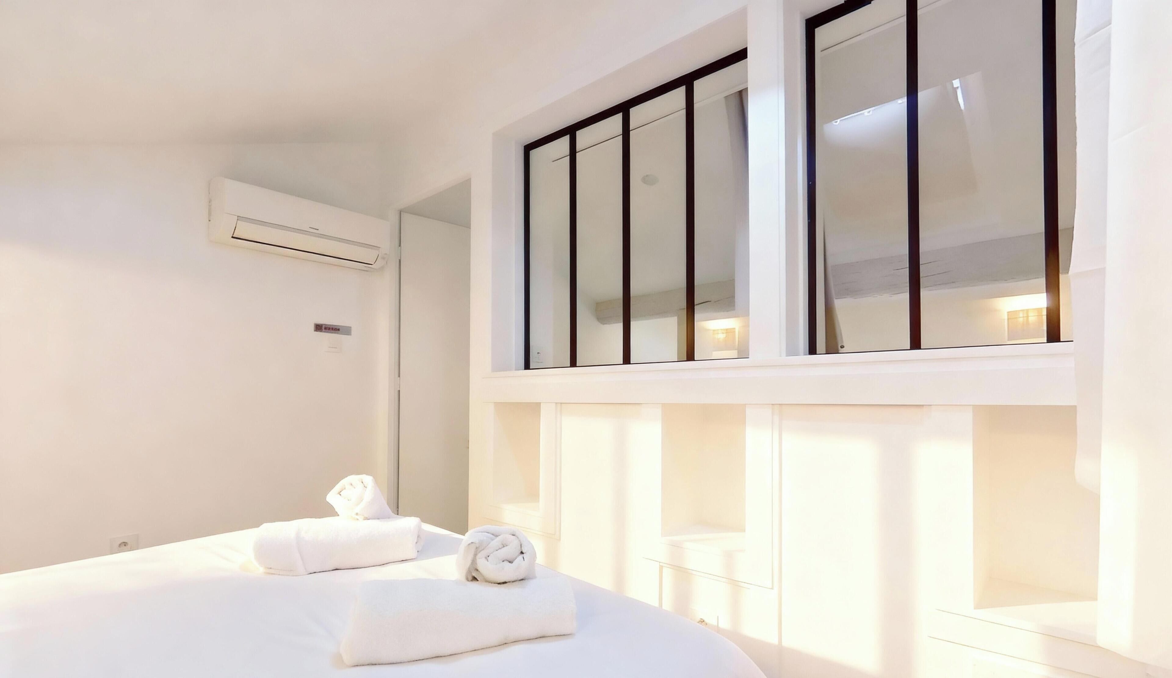 A hotel room with a large bed, a window with blinds, an air conditioning unit, and a small bedside table with a towel.