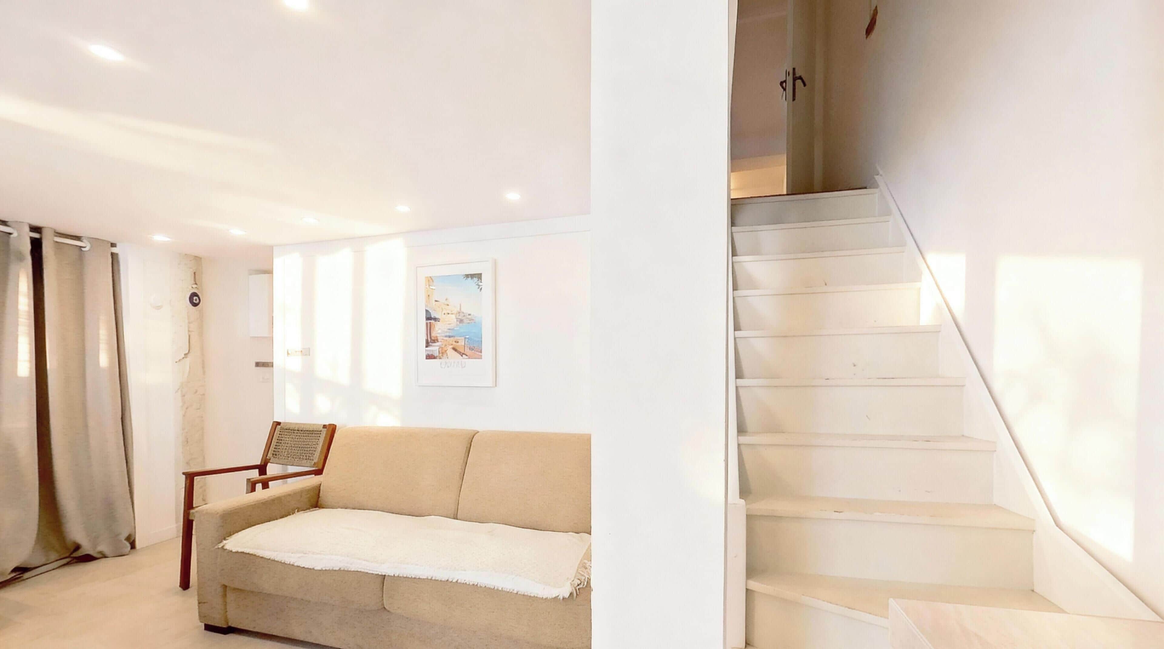 A modern interior with a beige sofa, a wooden chair, a framed picture on the wall, and a staircase.