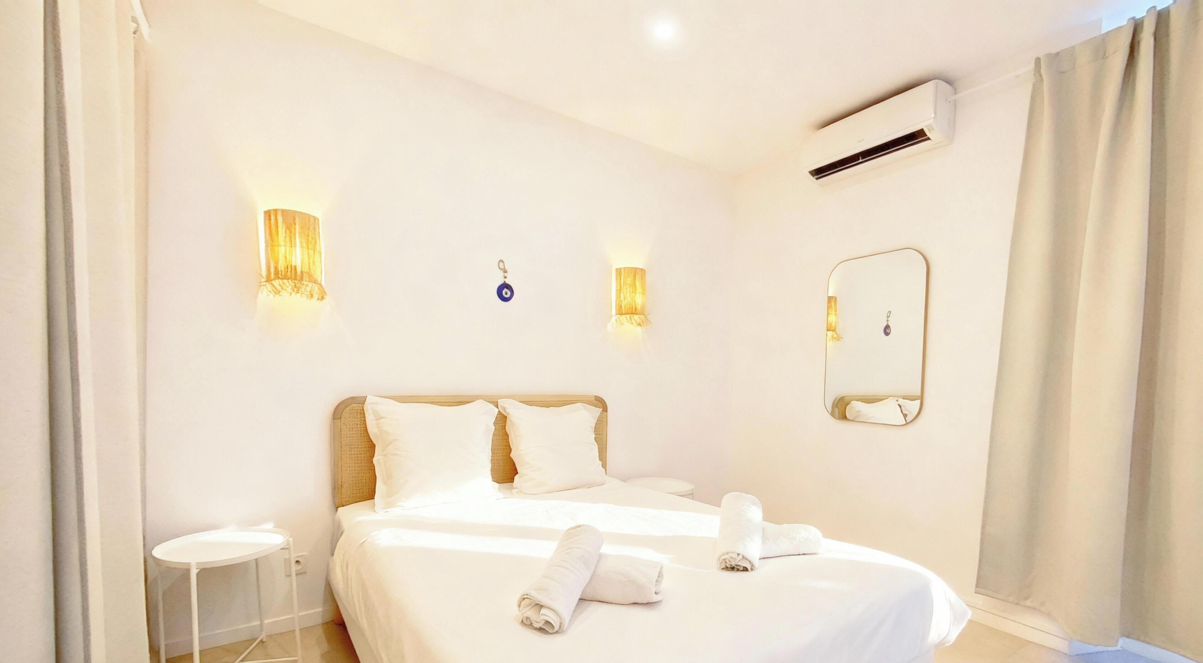 Quarto de hotel com cama, abajures, ar condicionado, espelho e uma mesinha.
