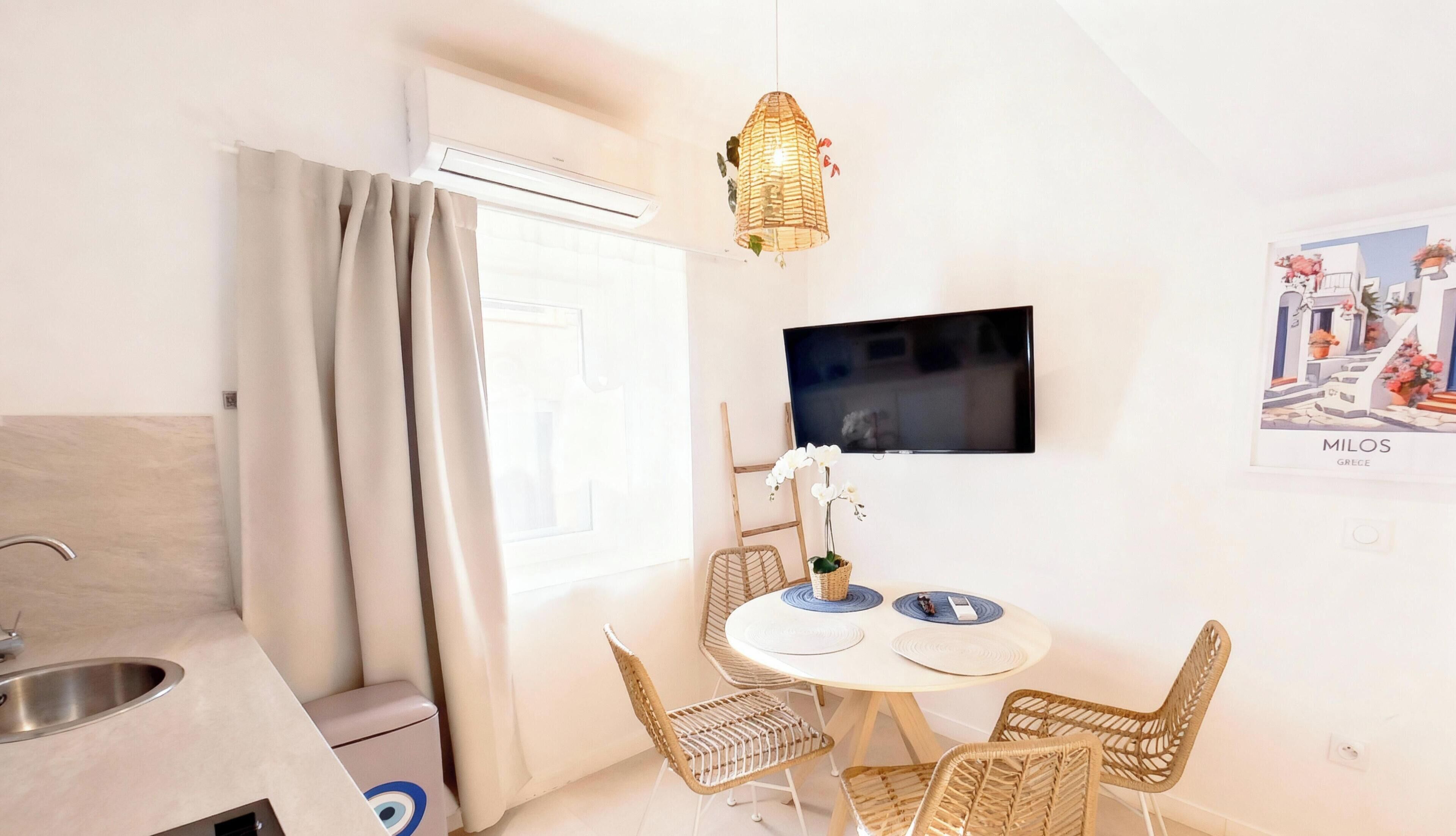 Une petite pièce lumineuse comprenant un coin cuisine, une table à manger avec des chaises en osier, un téléviseur à écran plat fixé au mur et une affiche représentant une ville grecque.