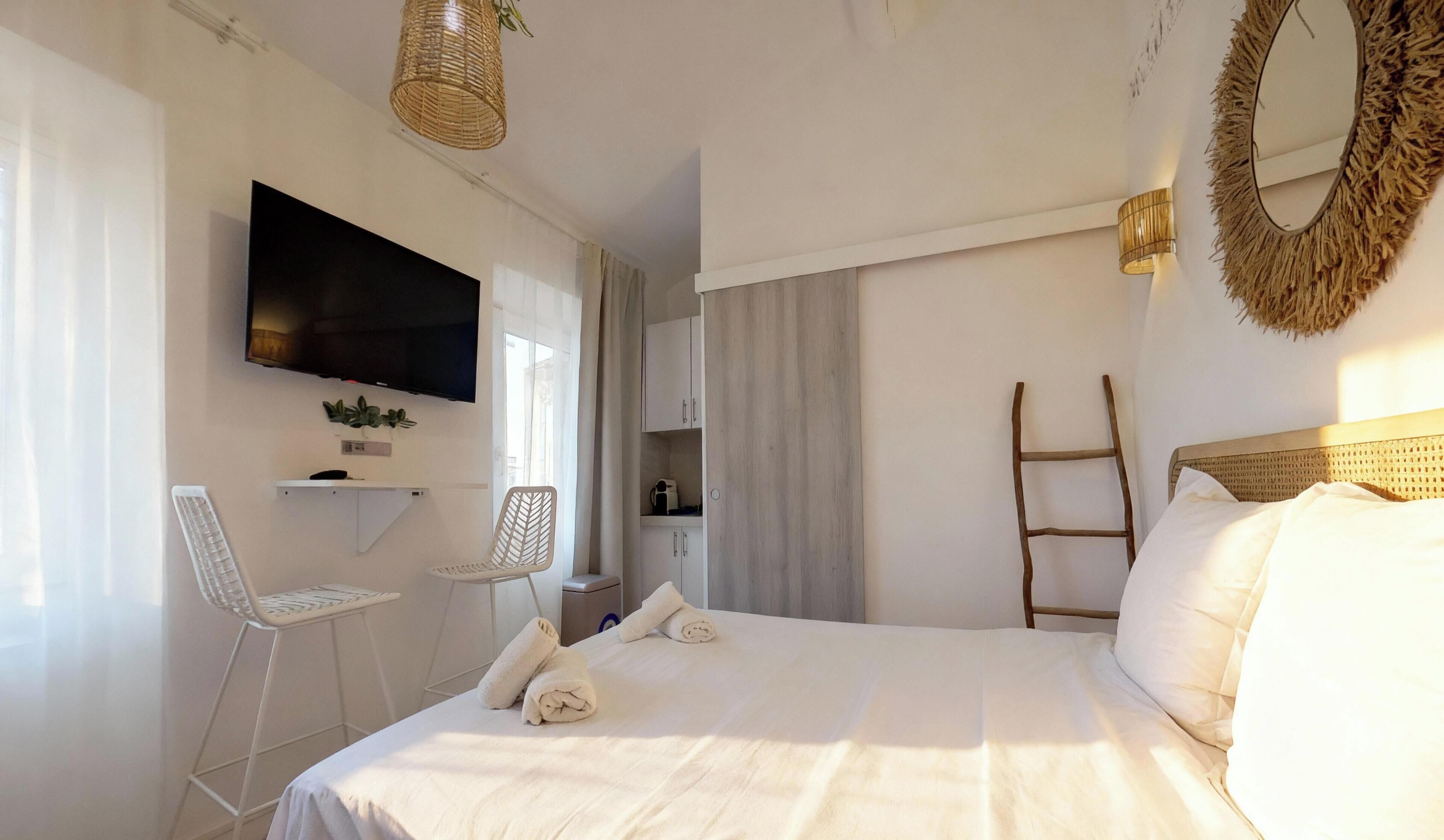 A hotel room with a bed, a TV mounted on the wall, a small desk with a chair, a ladder, and a mirror with a natural frame.