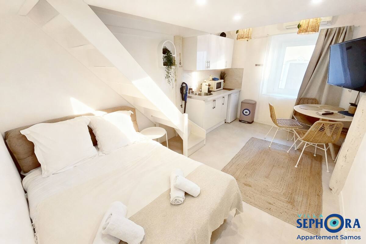 A modern hotel room with a bed, a small kitchenette, a dining area, and a TV.