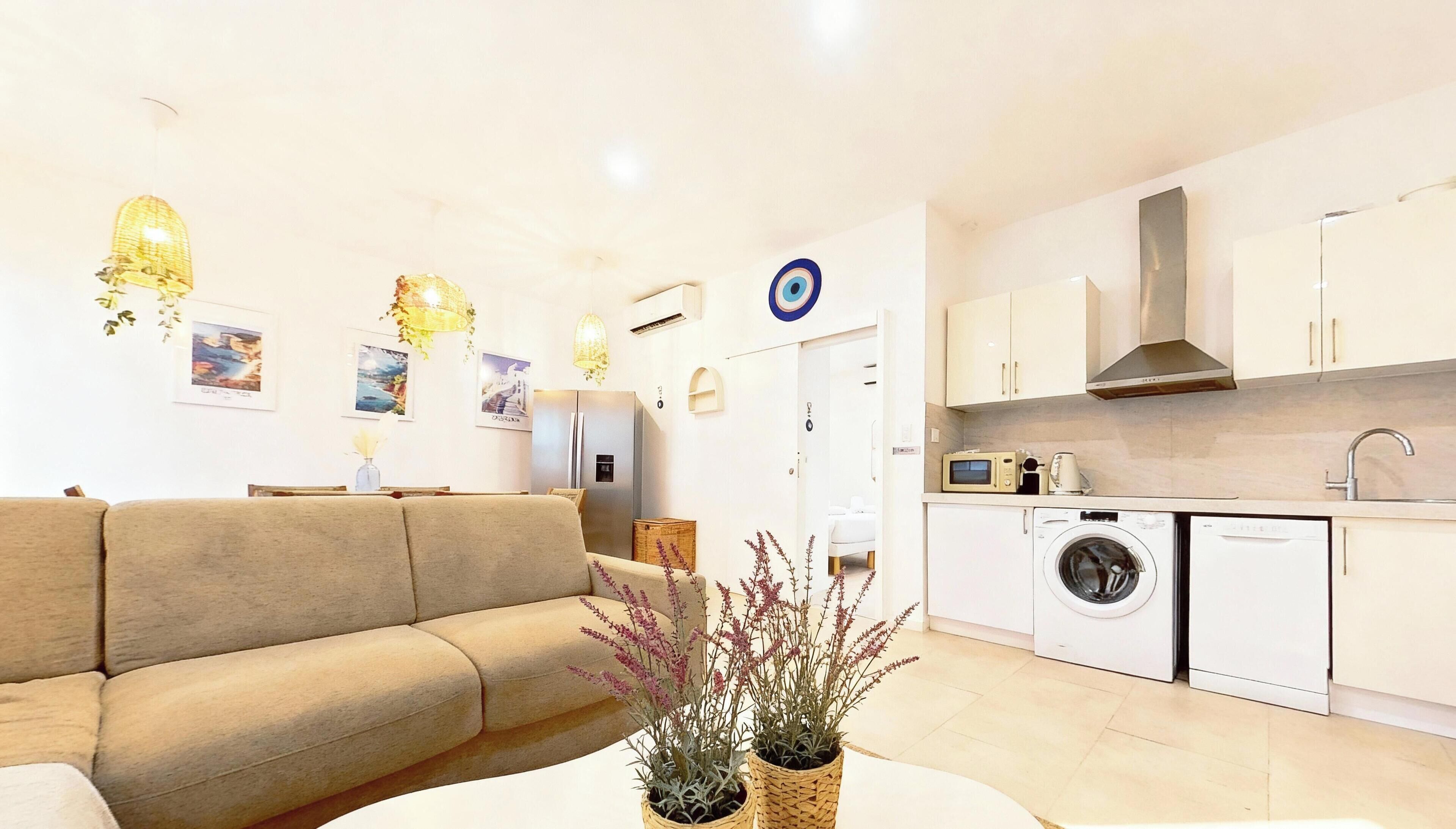 A modern living room with a beige sofa, a dining table, and a kitchen area with white cabinets, a washing machine, and a microwave.