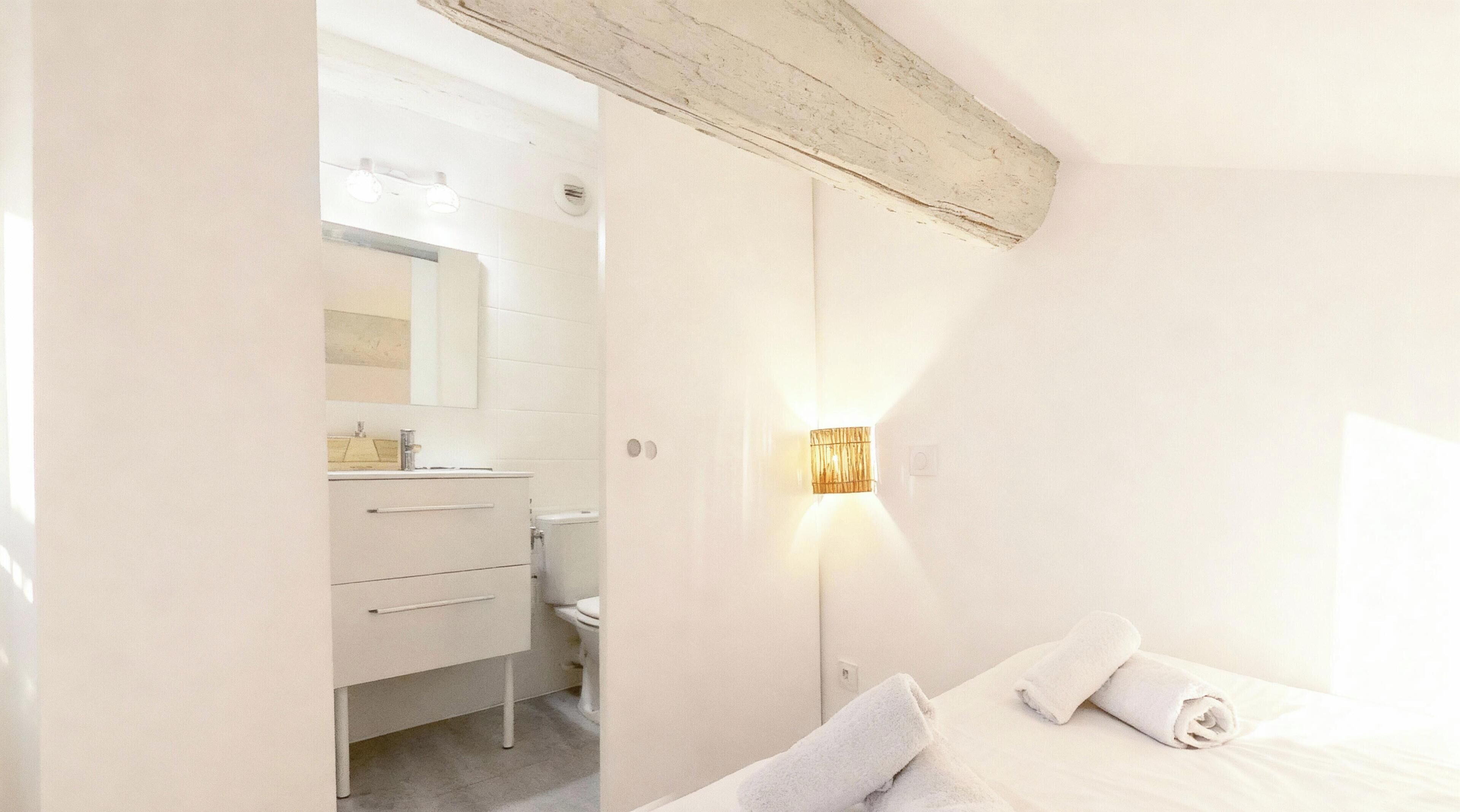 Una camera da letto con un letto, un comodino, una lampada a parete e un bagno con lavandino e specchio.