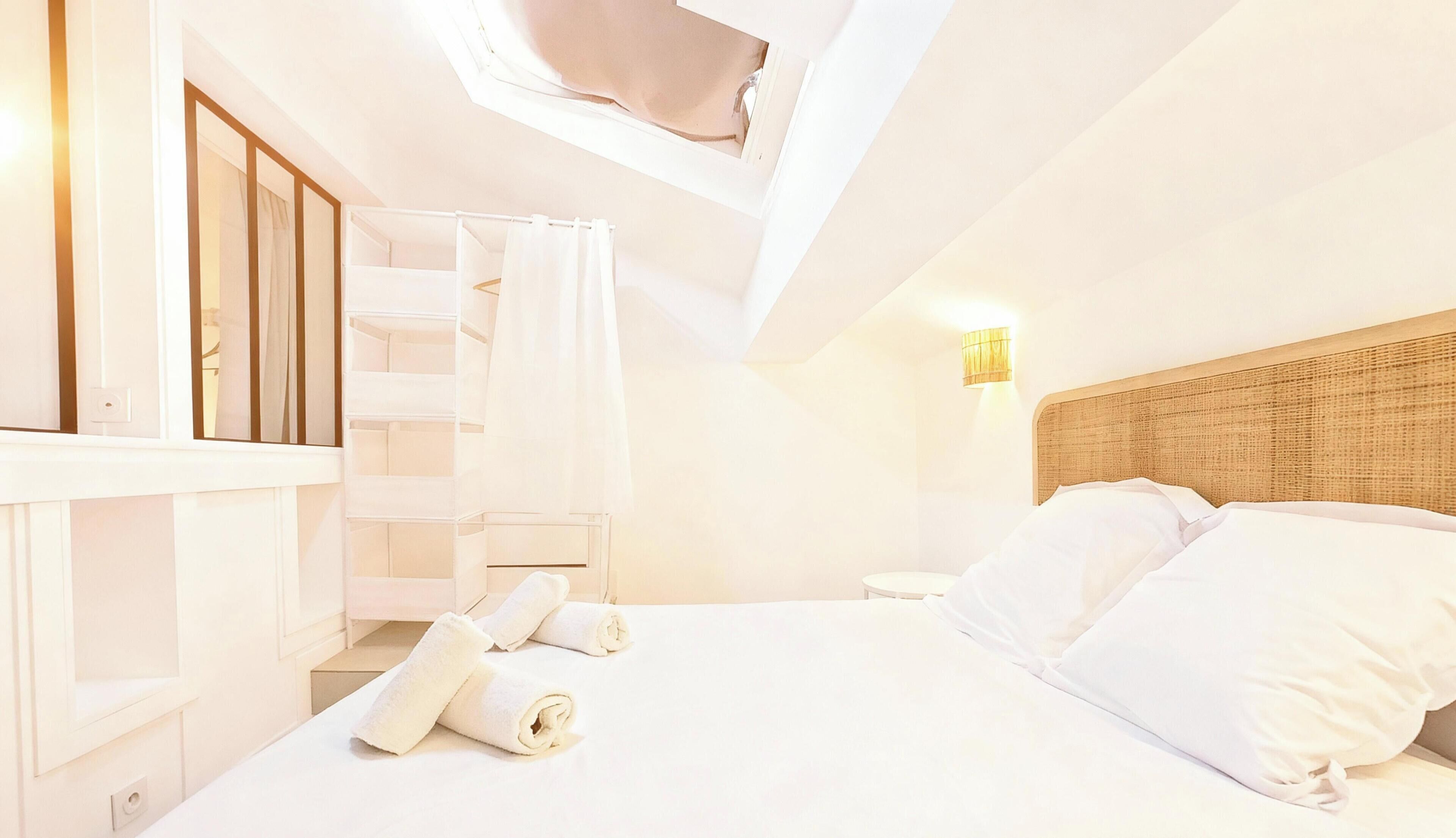 Un dormitorio con tragaluz, cama con ropa de cama blanca, cabecera de madera y lámpara de pared.