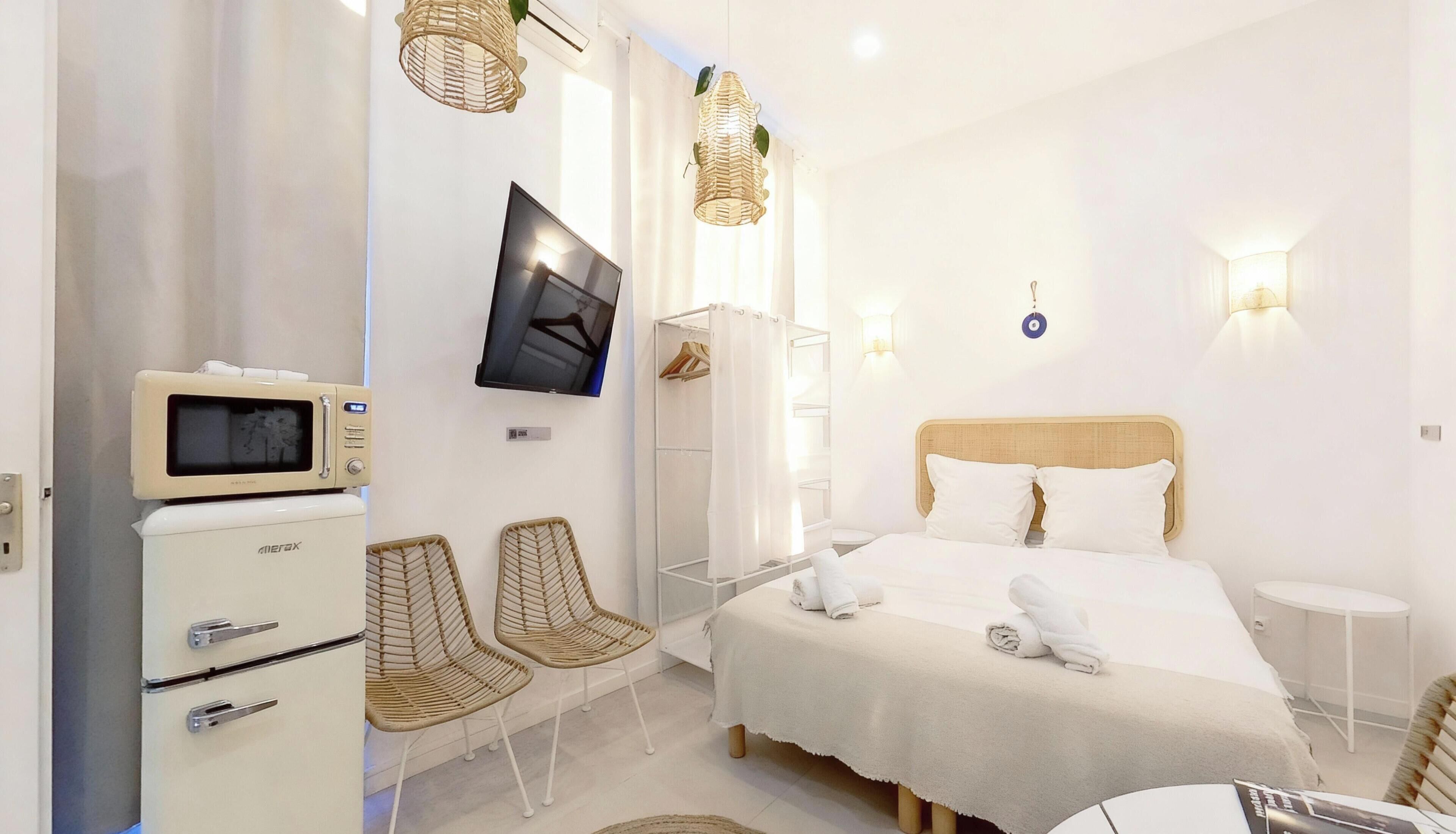 A modern hotel room with a bed, a TV, a minibar, wicker chairs, and a shower area.