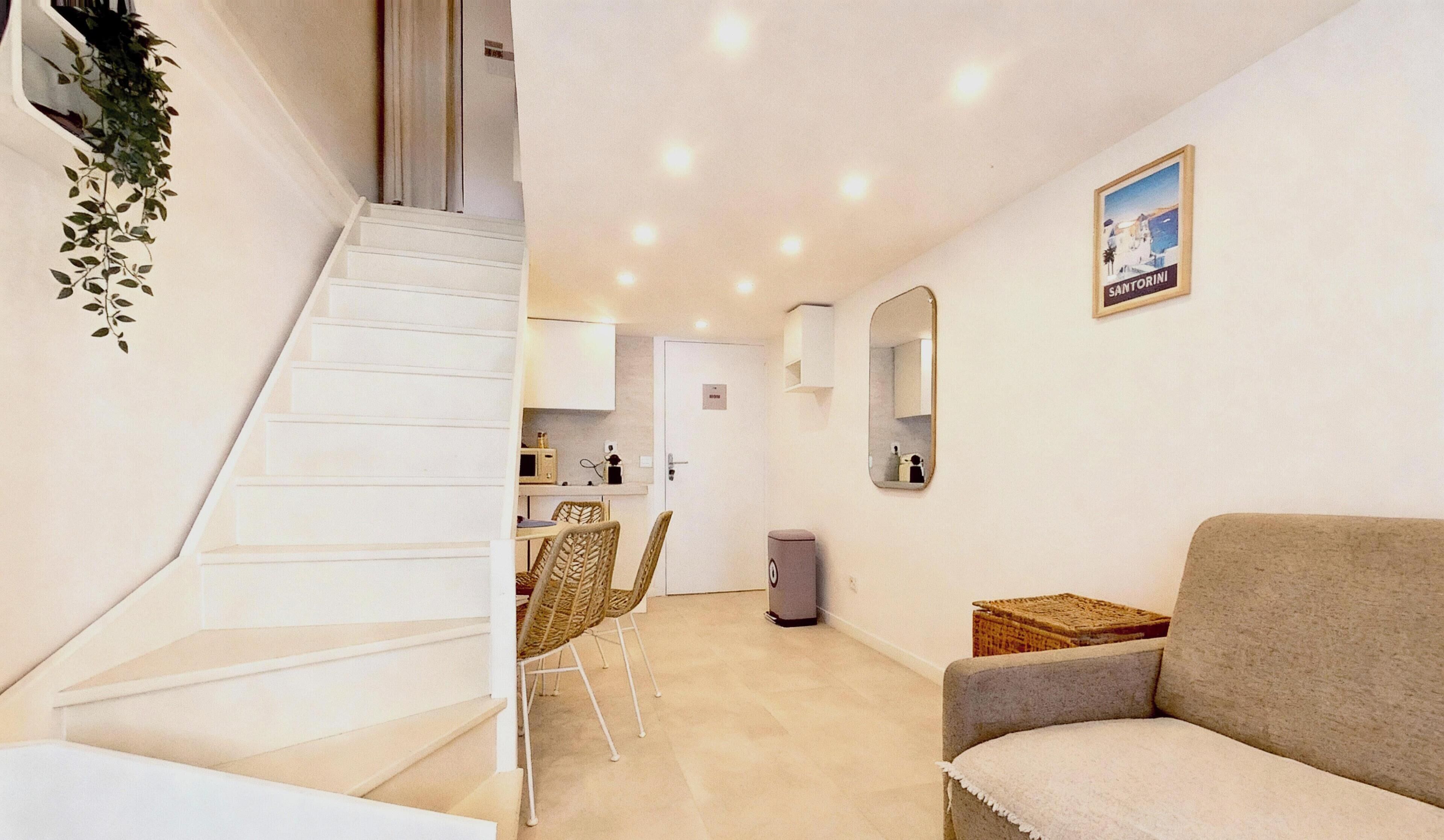 A modern apartment with a staircase leading to a kitchen, a dining area, and a living room with a sofa and a mirror on the wall.