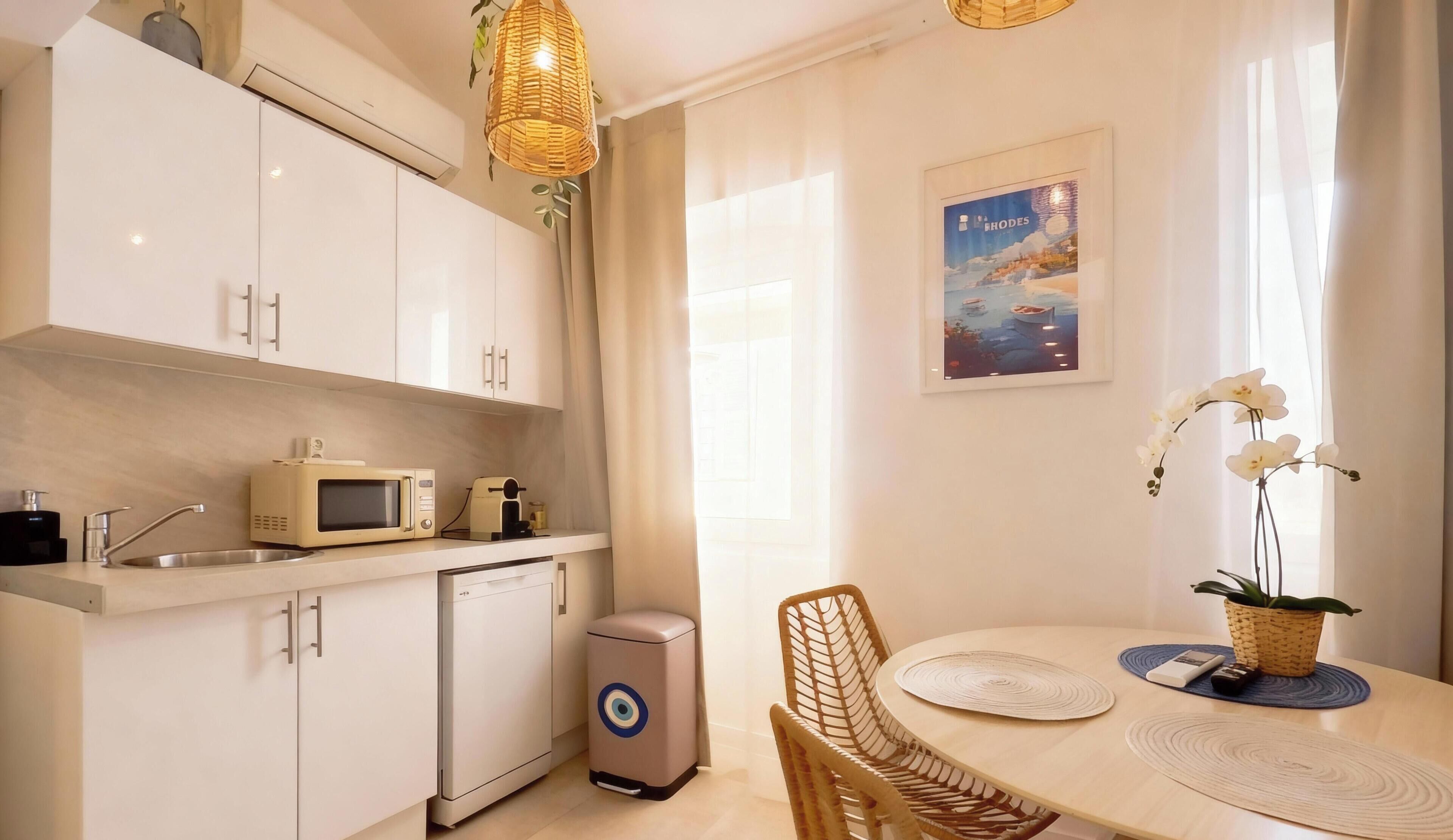 Una cocina moderna con armarios blancos, microondas, tostadora y lavavajillas. Hay una mesa de comedor con una silla, una planta decorativa y un cuadro en la pared.