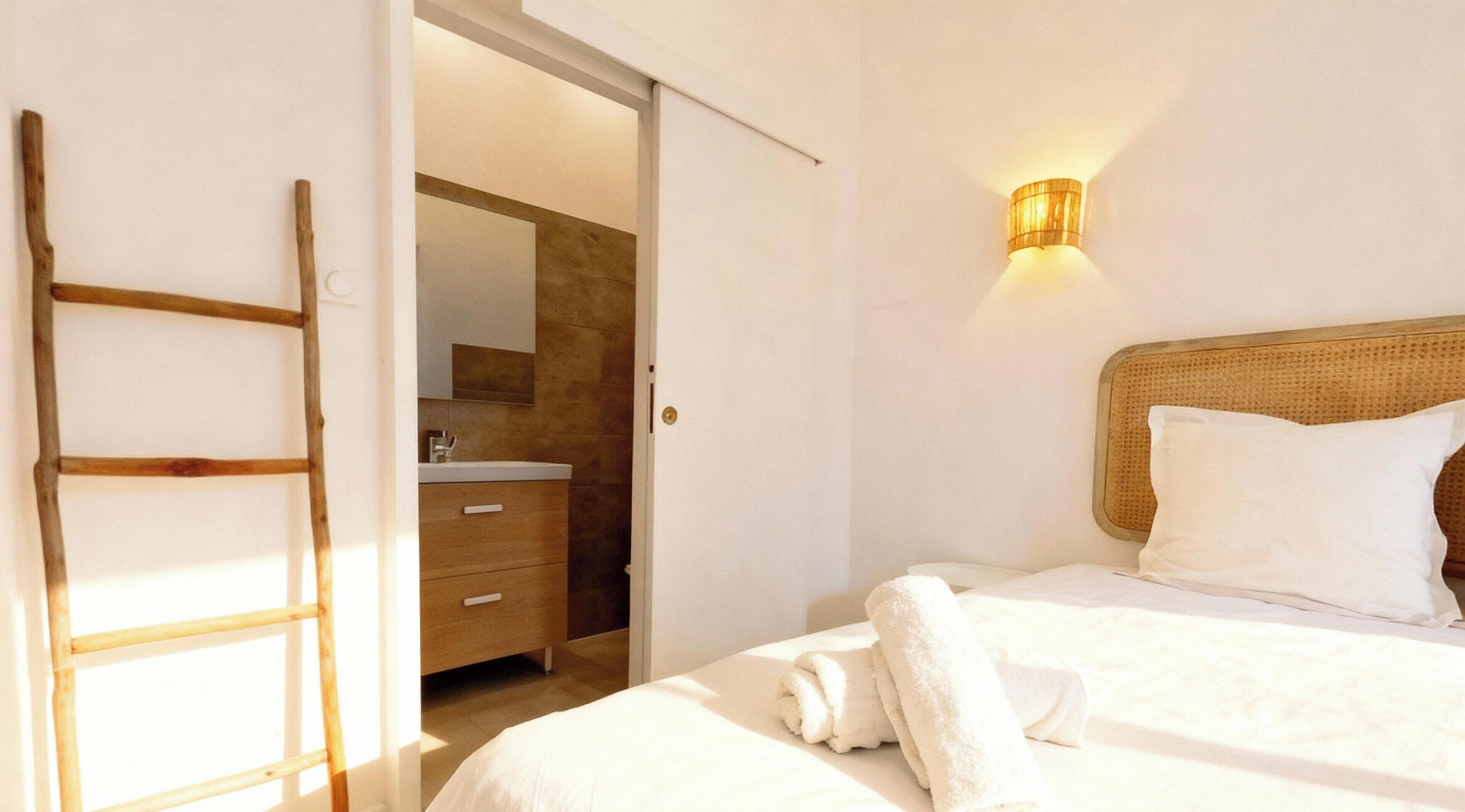 Un dormitorio con cama, una lámpara de noche, una escalera de madera y un baño visible a través de una puerta abierta.
