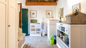 Un pasillo estrecho con suelo de madera, techo de piedra y una puerta verde. En las paredes hay cuadros enmarcados, un armario blanco con cajones y una cesta de mimbre sobre una encimera.