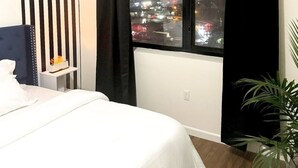 A hotel room with a bed, a nightstand, a window with black curtains, and a view of a cityscape at night. - Posh 2BR/2BA King Bed! Free Parking! 30 Min to NYC

 (East Orange)