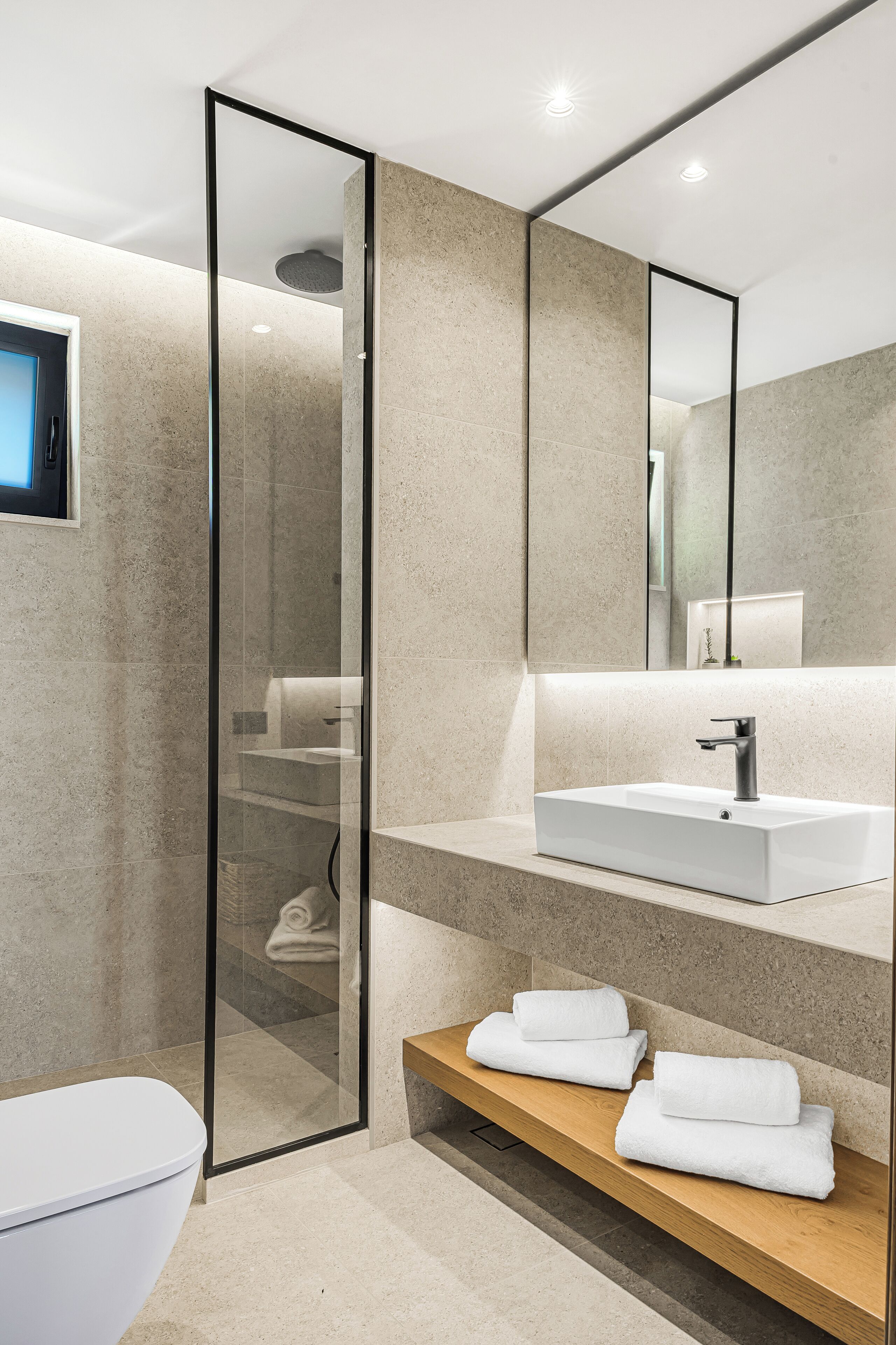 Un baño moderno con cabina de ducha de vidrio, un lavabo blanco y un estante de madera con toallas.