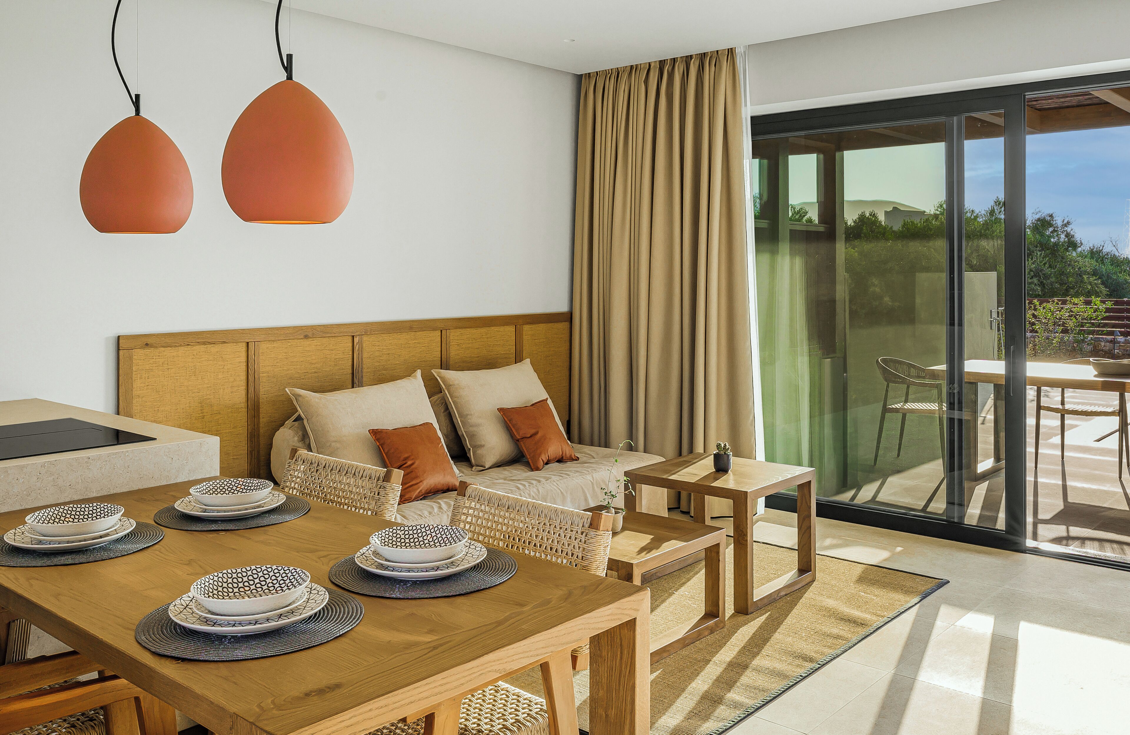 A modern hotel room with a wooden dining table set for a meal, a sofa with cushions, a small side table, and a large sliding glass door leading to an outdoor area.
