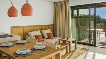 A modern hotel room with a wooden dining table set for a meal, a sofa with cushions, a small side table, and a large sliding glass door leading to an outdoor area.