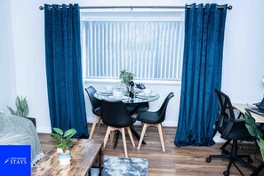A dining area with a round table, black chairs, and a window with blinds. - 3 Bed House Sleeps 7 With Garden and Parking (Walsall)