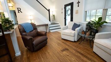 Un salon avec un fauteuil en cuir marron, un fauteuil blanc et une petite table sur laquelle est posée une plante. On aperçoit un escalier avec une rampe au fond.
