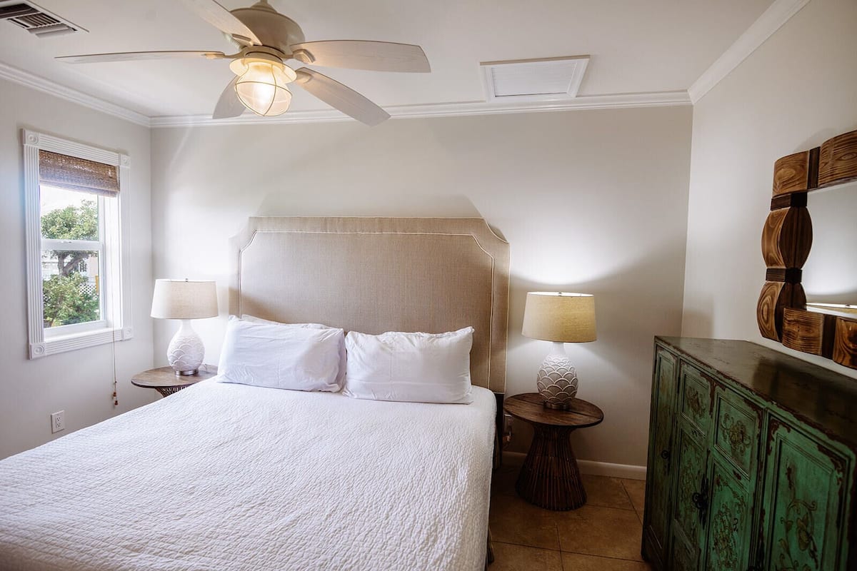 A bedroom with a bed, two bedside lamps, a ceiling fan, a window with a view, and a green dresser.