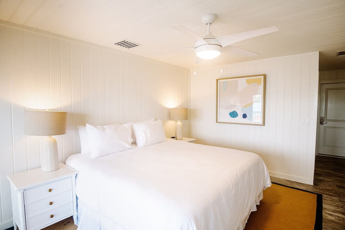 A white bedroom with a bed, two bedside tables with lamps, a ceiling fan, and a framed abstract painting on the wall.