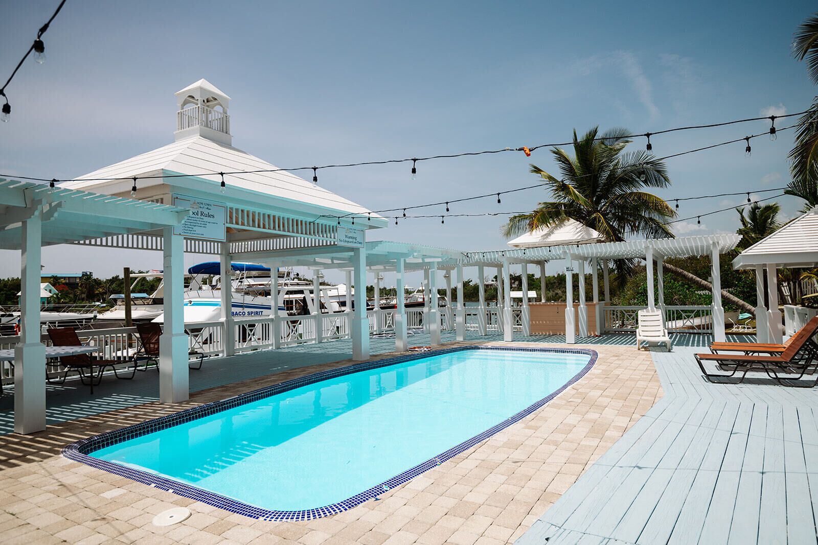 Un espace piscine avec une piscine à l’eau d’un bleu limpide, des pergolas blanches, des chaises longues et un bateau amarré au fond.