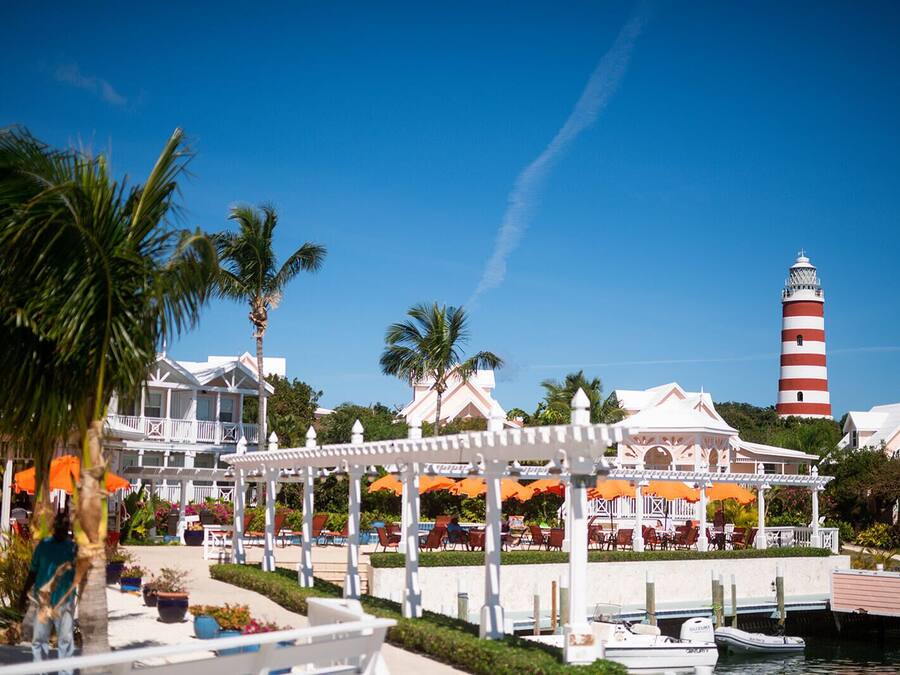 Un resort con un pabellón blanco, mobiliario exterior, un puerto deportivo y un faro al fondo.