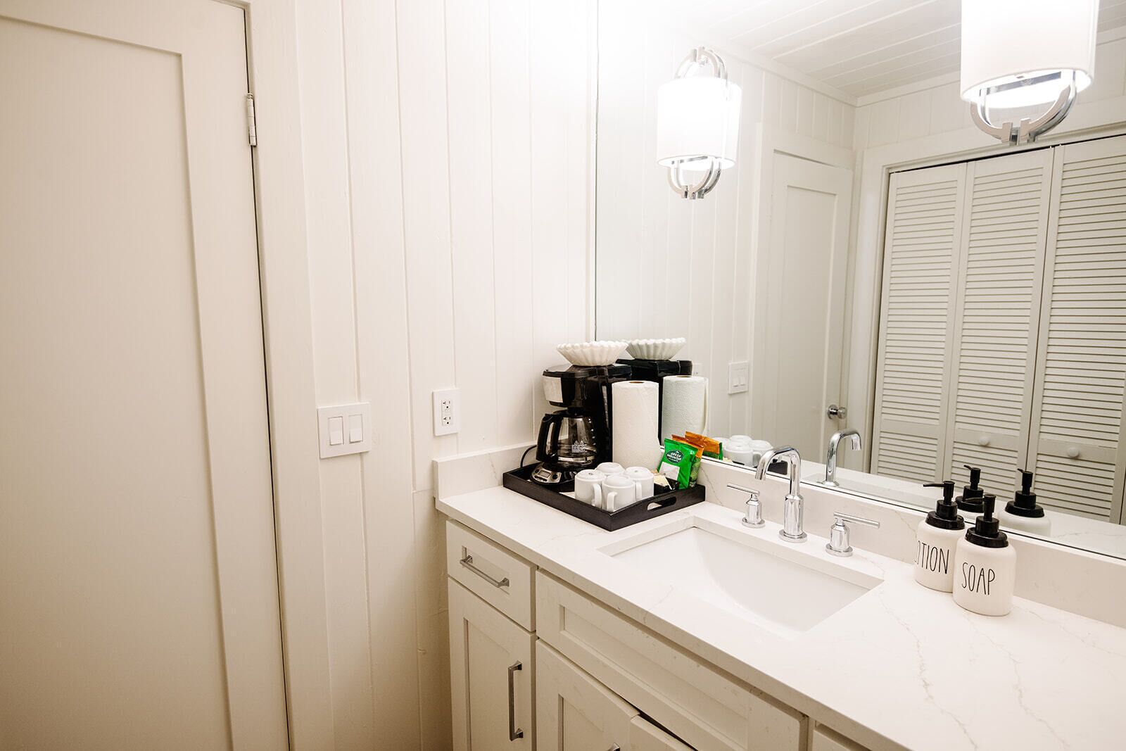 A hotel bathroom with a white vanity, a coffee maker, a soap dispenser, and a mirror.