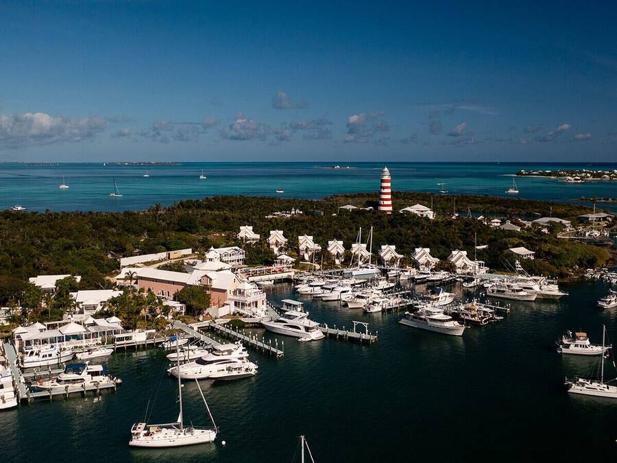 An aerial view of a marina with numerous boats docked, a lighthouse, and a cluster of buildings with a hilly backdrop.