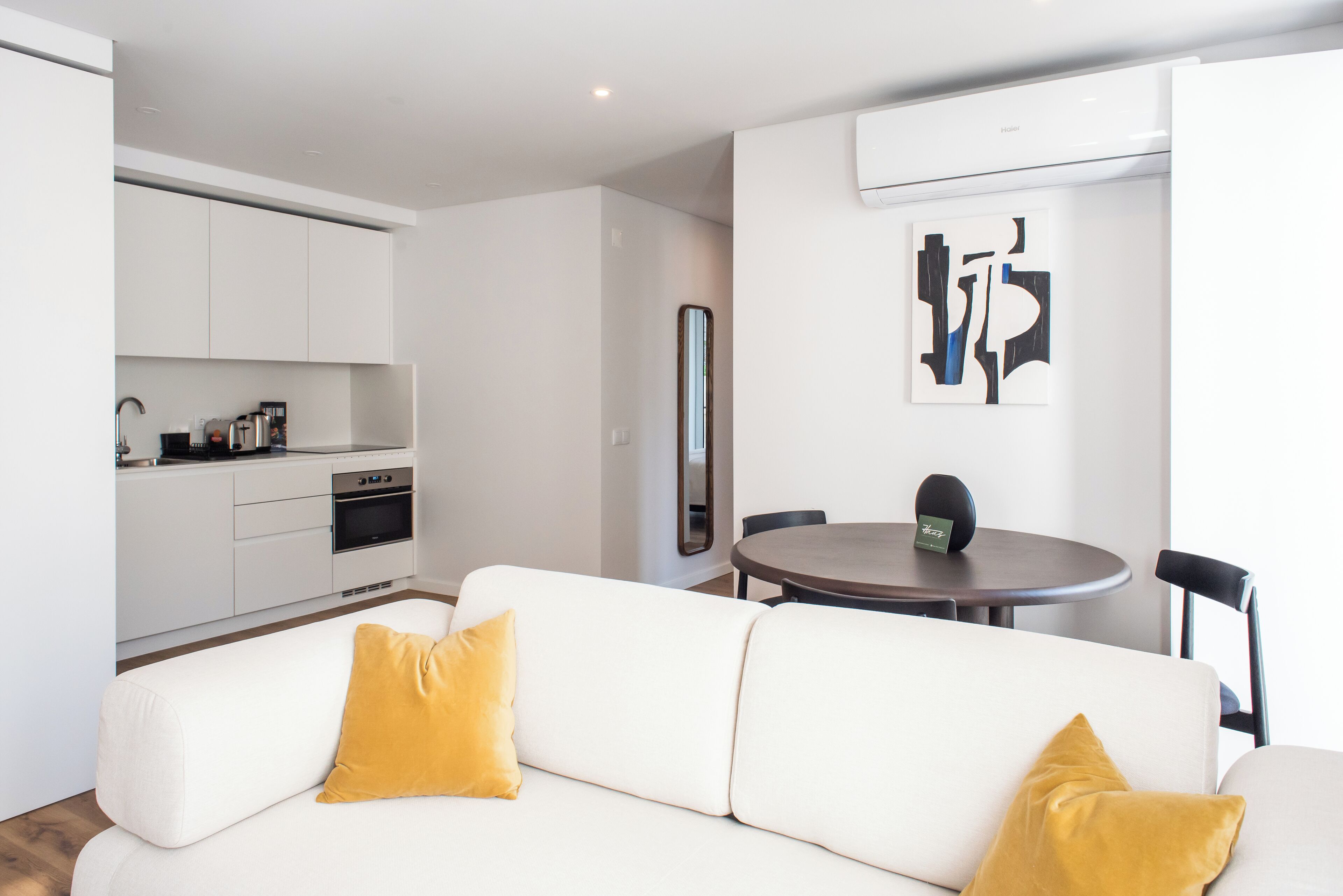 Una sala de estar moderna con un sofá blanco, dos cojines color mostaza, una mesa de comedor con una silla, y una zona de cocina con armarios y electrodomésticos blancos.