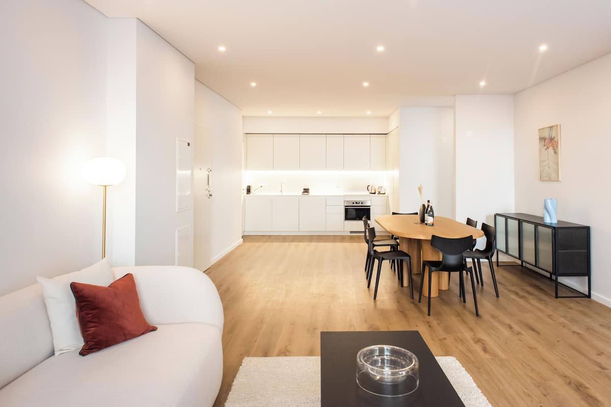 Un salon moderne avec un canapé blanc, un coussin rouge et une table basse surmontée d’un saladier en verre. On y trouve également un coin repas avec une table et des chaises en bois, et une cuisine à l’arrière-plan.