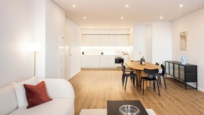 Un salon moderne avec un canapé blanc, un coussin rouge et une table basse surmontée d’un saladier en verre. On y trouve également un coin repas avec une table et des chaises en bois, et une cuisine à l’arrière-plan.