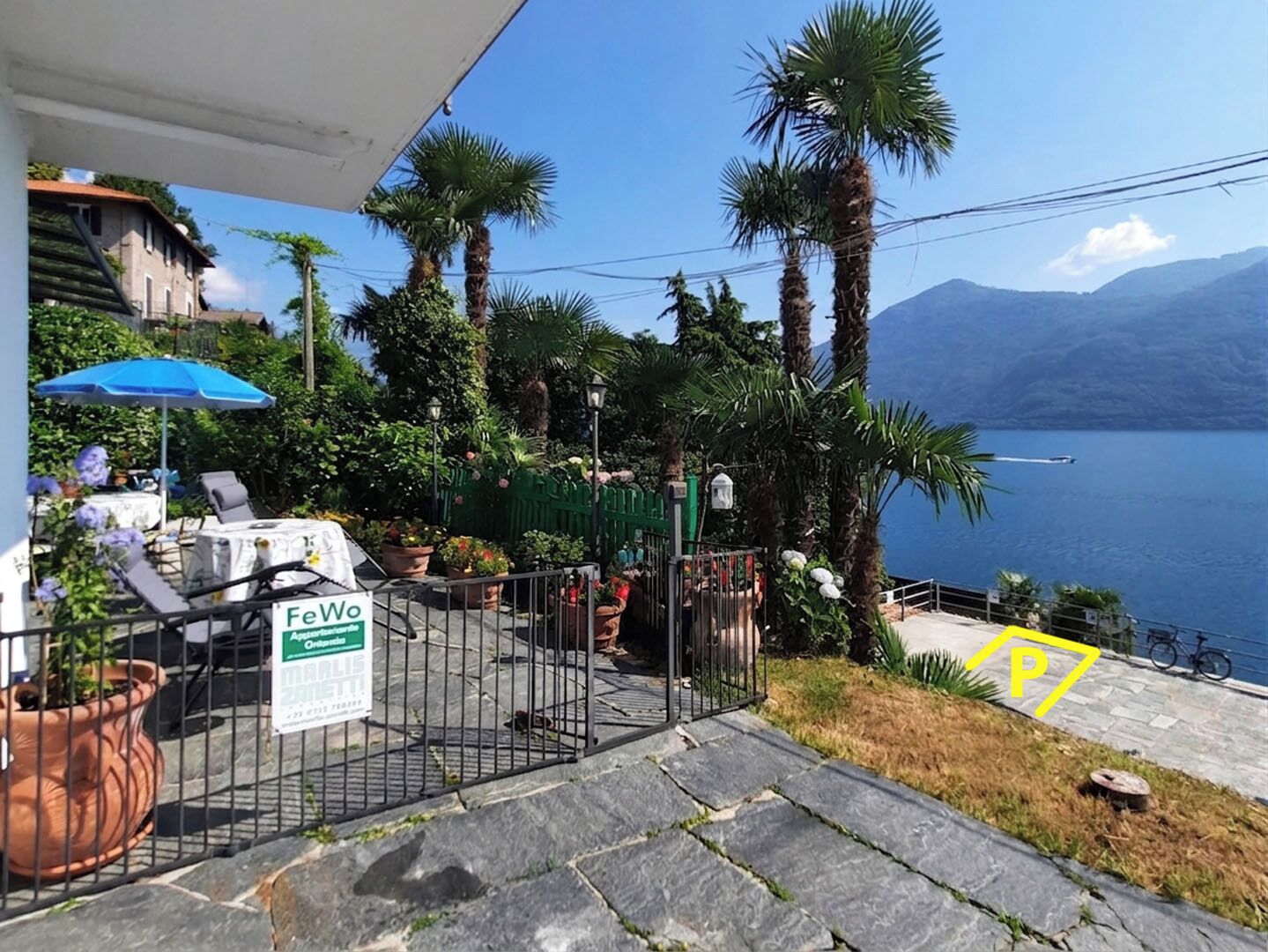 Una terraza con asientos exteriores, un cartel que dice 'FeWo' y vistas a un lago con montañas al fondo.