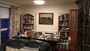 Comedor con una mesa preparada para dos personas, rodeado de estanterías llenas de libros. En la pared hay un cuadro de caballos, y una ventana con cortinas a la izquierda.