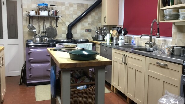 Una cocina con estufa, pileta y diversos utensilios. Se observa una mesa de madera con una olla encima.