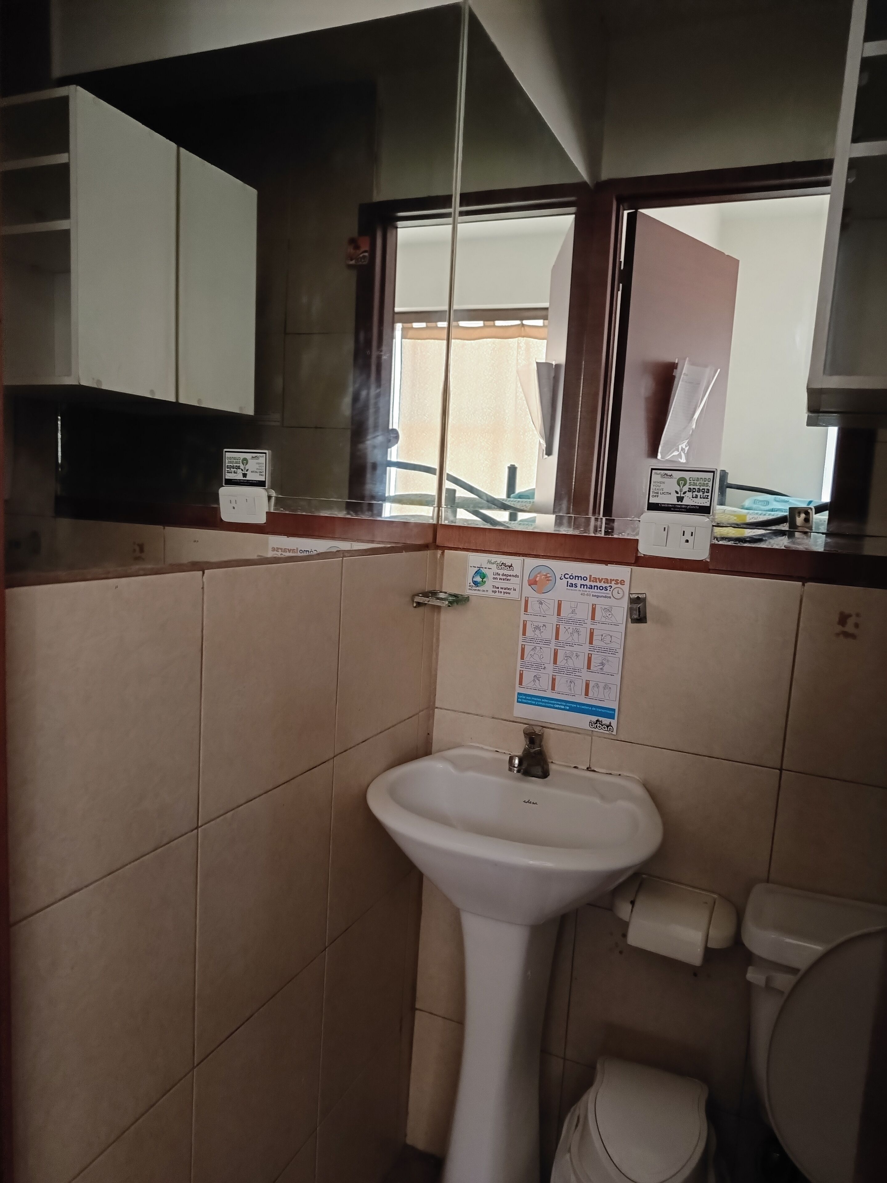 Un baño con un lavamanos blanco, un espejo y un inodoro. Hay una ventana con vistas al exterior y un cartel en la pared.