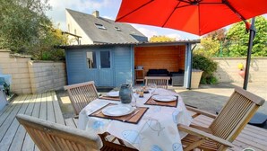 An outdoor dining area with a wooden deck, a table set for four, and a red umbrella providing shade. - Gîte la Crabière, 900m de la Mer, Animaux de Compagnie Bienvenue (SAINT CAST le guildo)