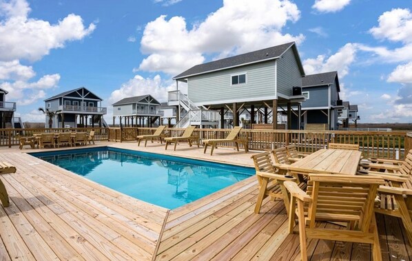 A wooden deck with a pool, wooden lounge chairs, and a view of houses in the background. - "Seas the Day" - Family Friendly Home Next to Pool and Playground (Bolivar Peninsula)