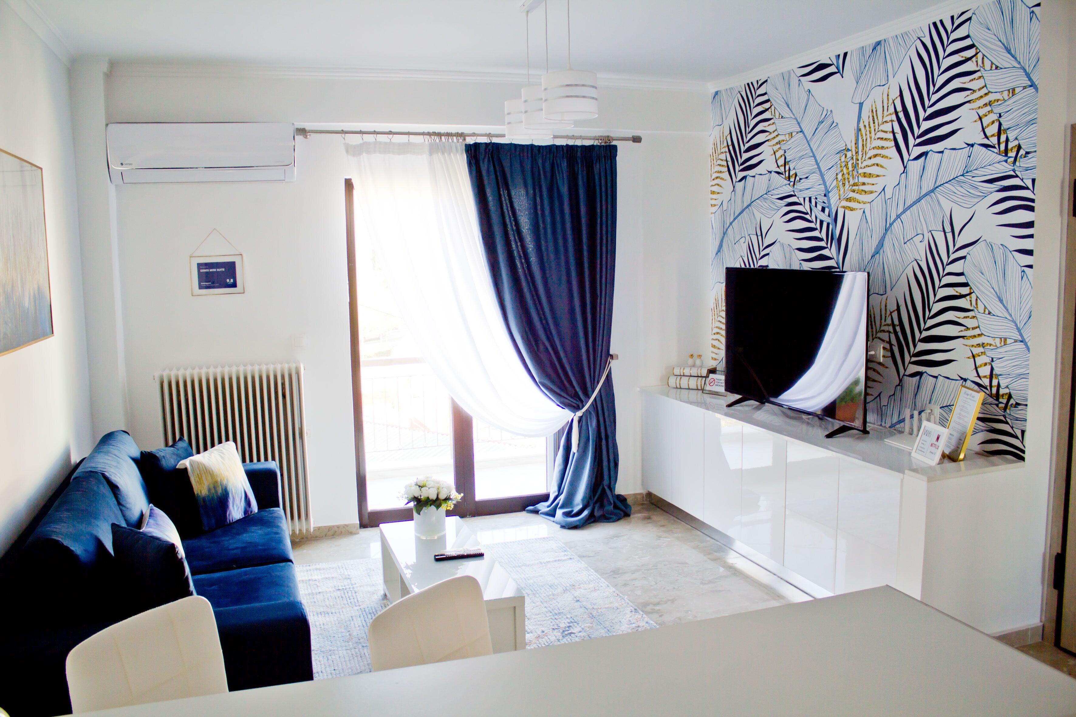 A modern living room with a blue sofa, white dining table, and a TV on a white cabinet.