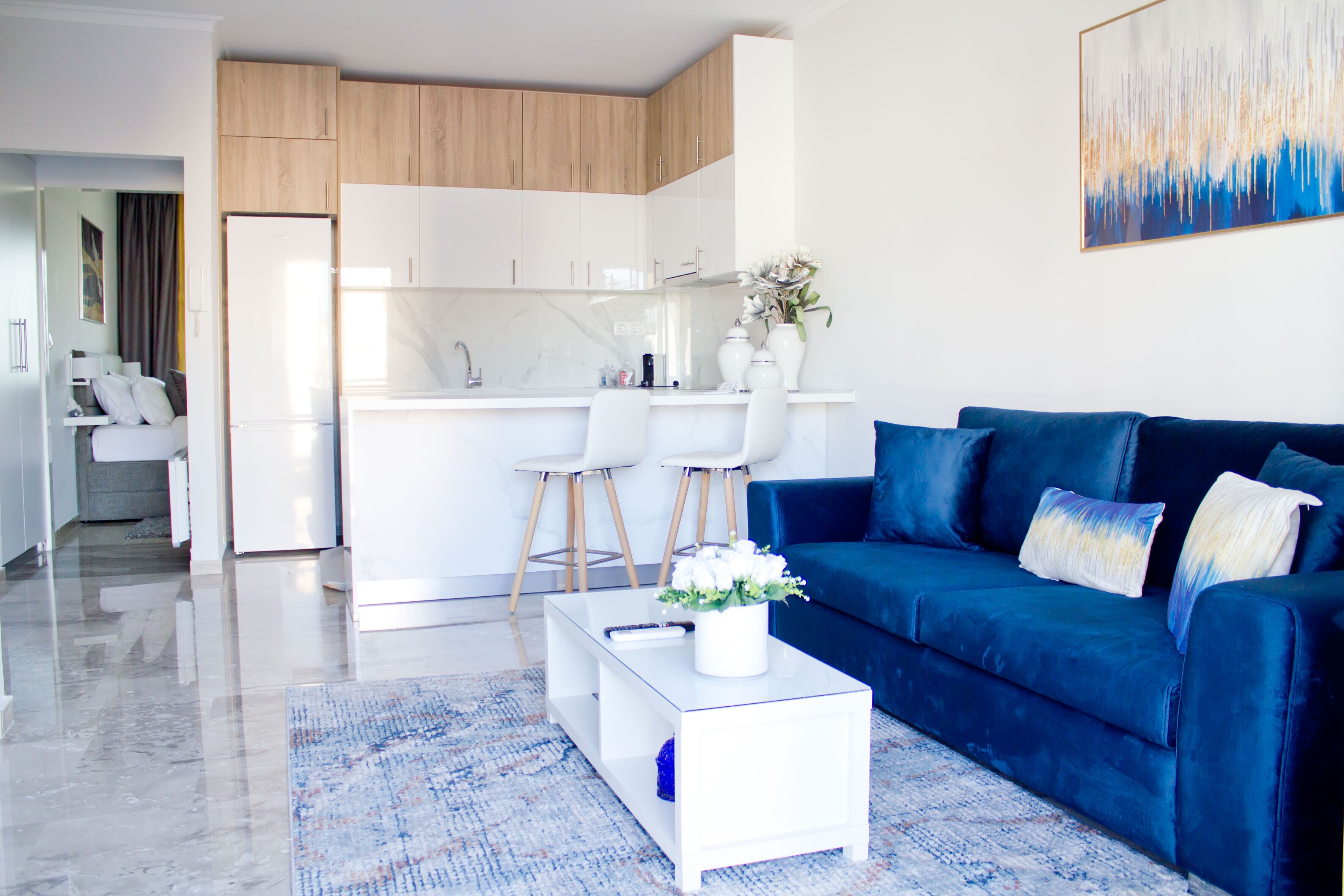 A modern living room with a blue sofa, a white coffee table, and a kitchen area with white cabinets and a marble countertop.