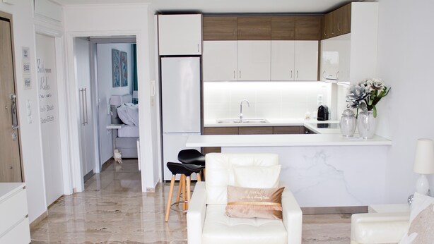 Una cocina moderna con armarios blancos y encimera de mármol. Hay un sofá blanco y una silla negra en la sala de estar.