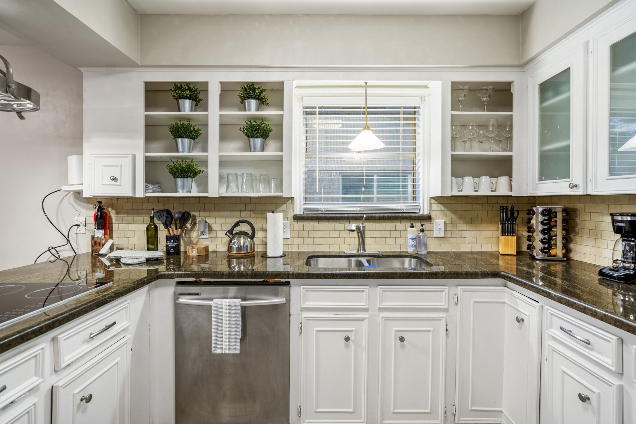 A modern kitchen with white cabinets, a granite countertop, a built-in oven, and a window with blinds.