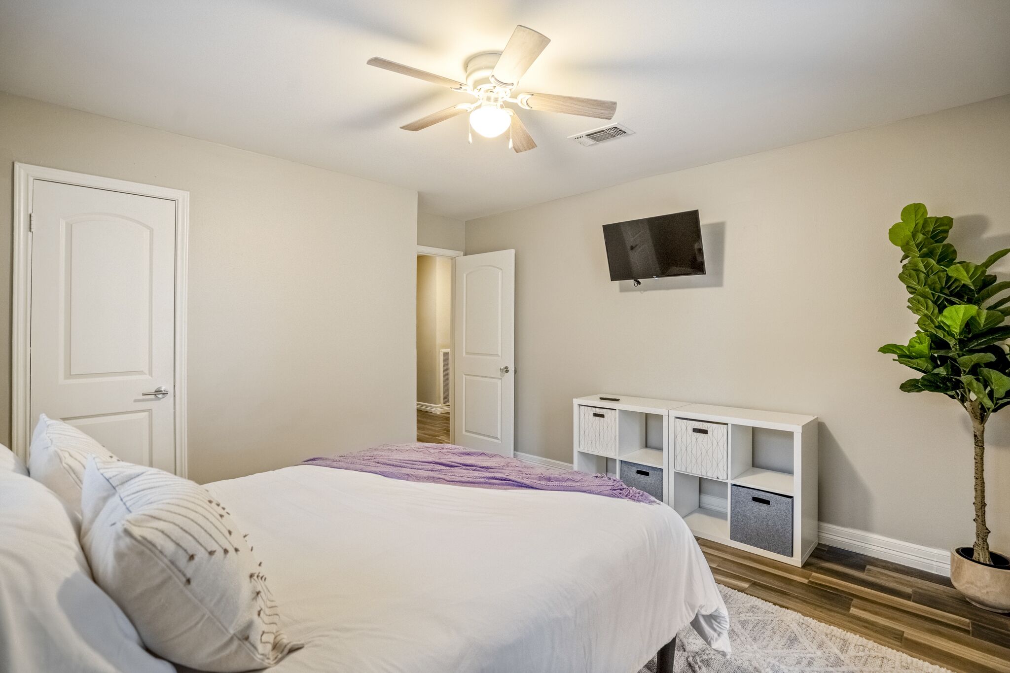 A bedroom with a bed, a ceiling fan, a TV on the wall, a plant, and a storage unit.