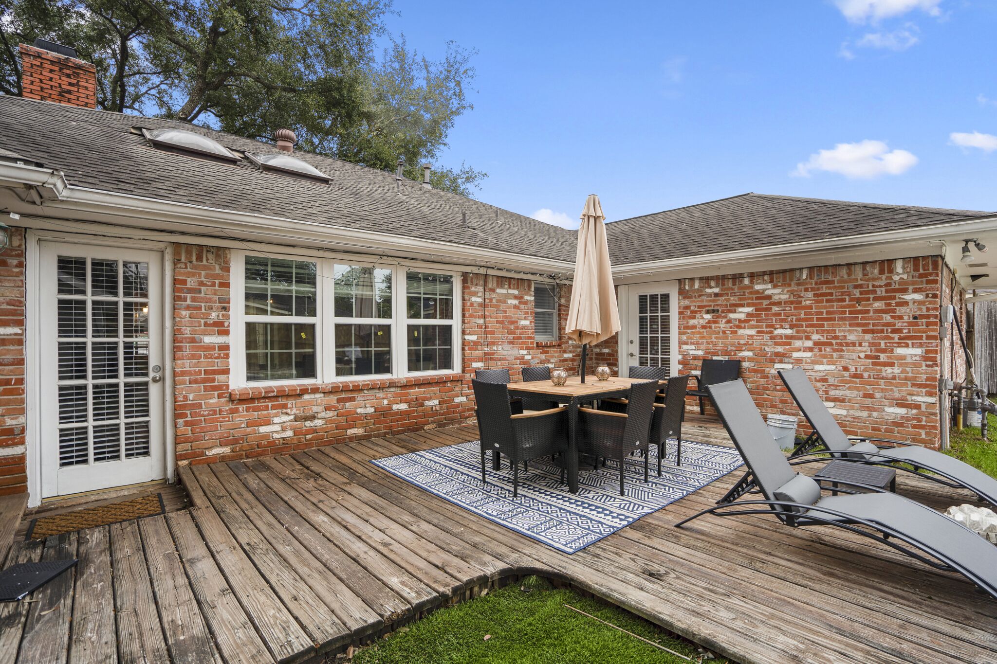 A backyard with a wooden deck, a brick house, a dining table with chairs, and a sun umbrella.