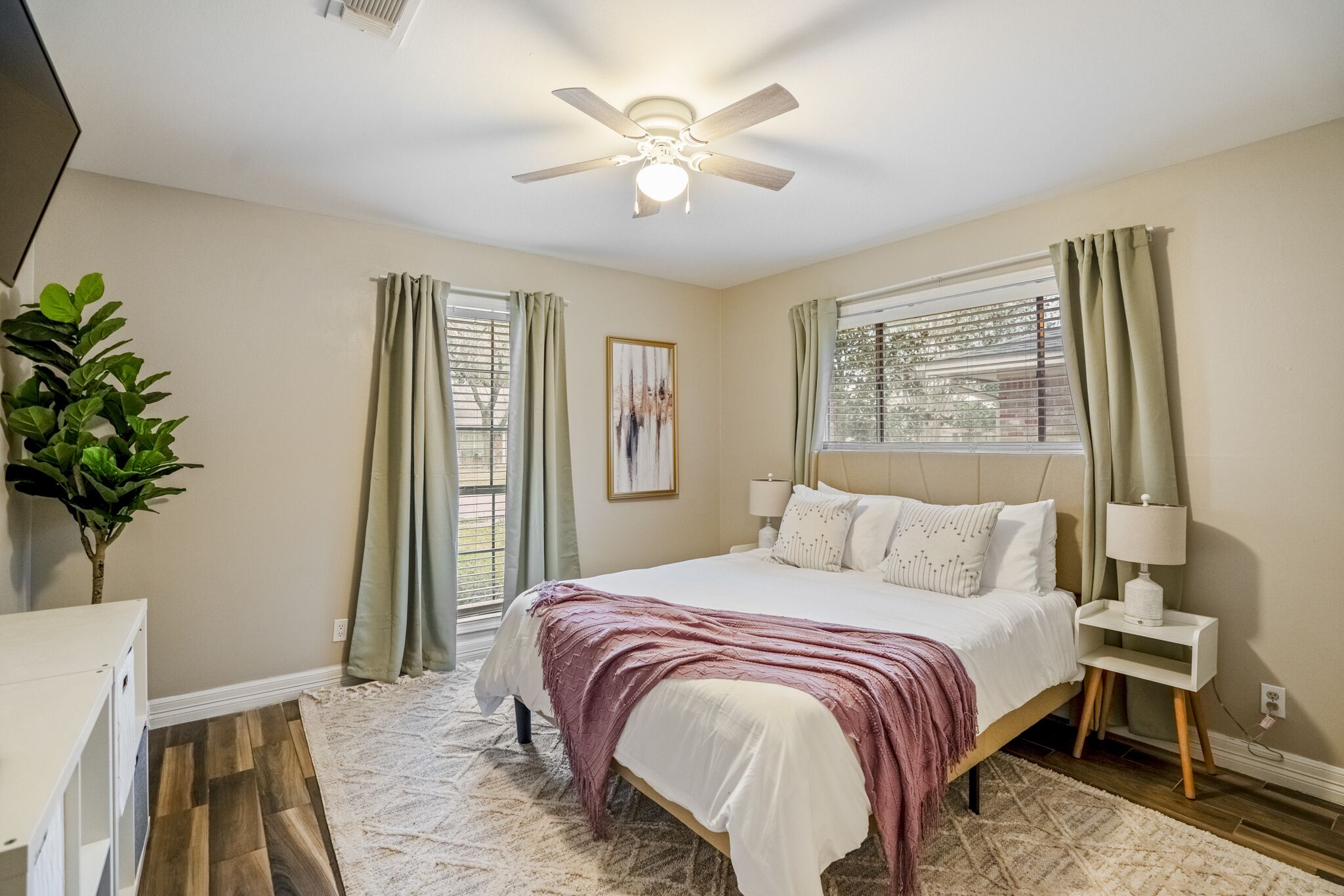 A bedroom with a bed, a ceiling fan, a window with curtains, a bedside table with a lamp, and a plant.