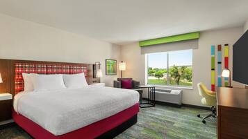 A hotel room with a large bed, a desk, a chair, a window with a view, and a wall with a colorful artwork.