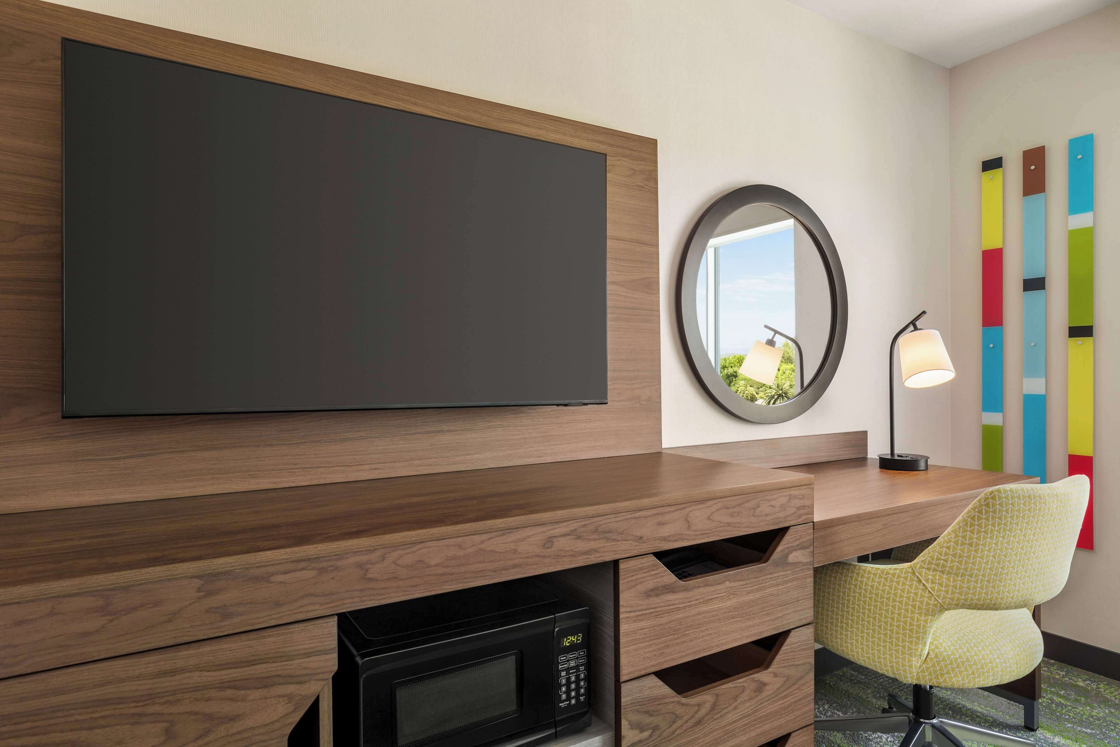 Une chambre d’hôtel moderne dotée d’un grand téléviseur à écran plat, d’un miroir rond, d’un bureau avec une lampe et d’œuvres d’art colorées accrochées au mur.