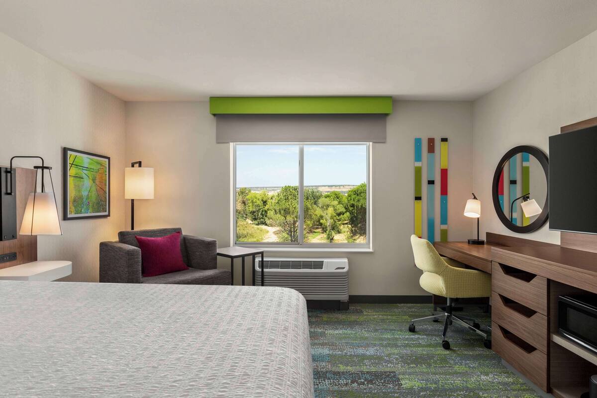 A hotel room with a large bed, a desk, a chair, a TV, a mirror, and a window with a view of greenery.