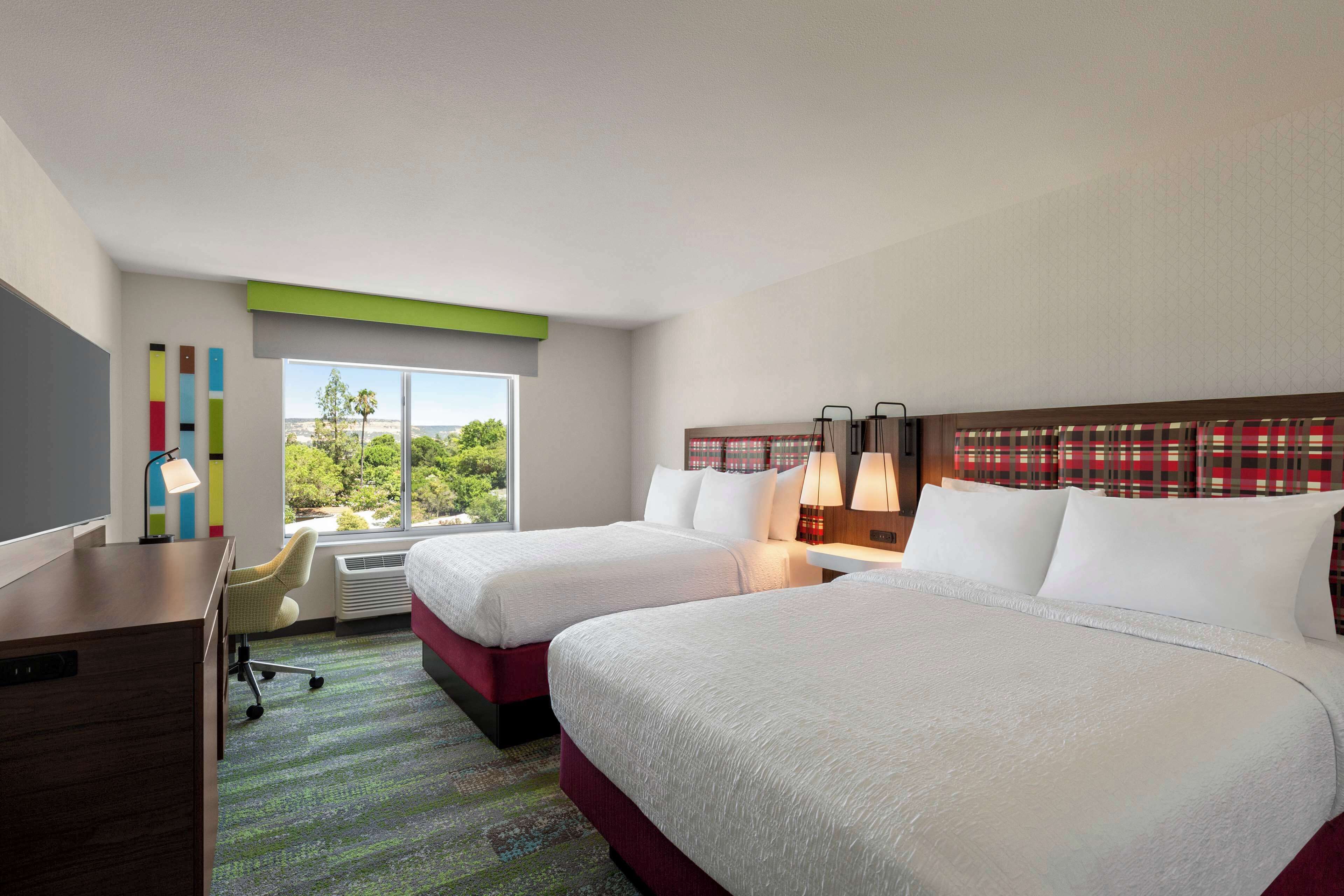 A hotel room with two beds, a desk, a chair, a window with a view of greenery, and a wall with a red and white checkered pattern.