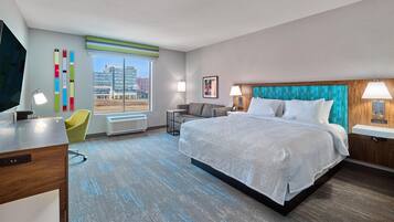 A modern hotel room with a large bed, a desk, a chair, and a view of the cityscape.