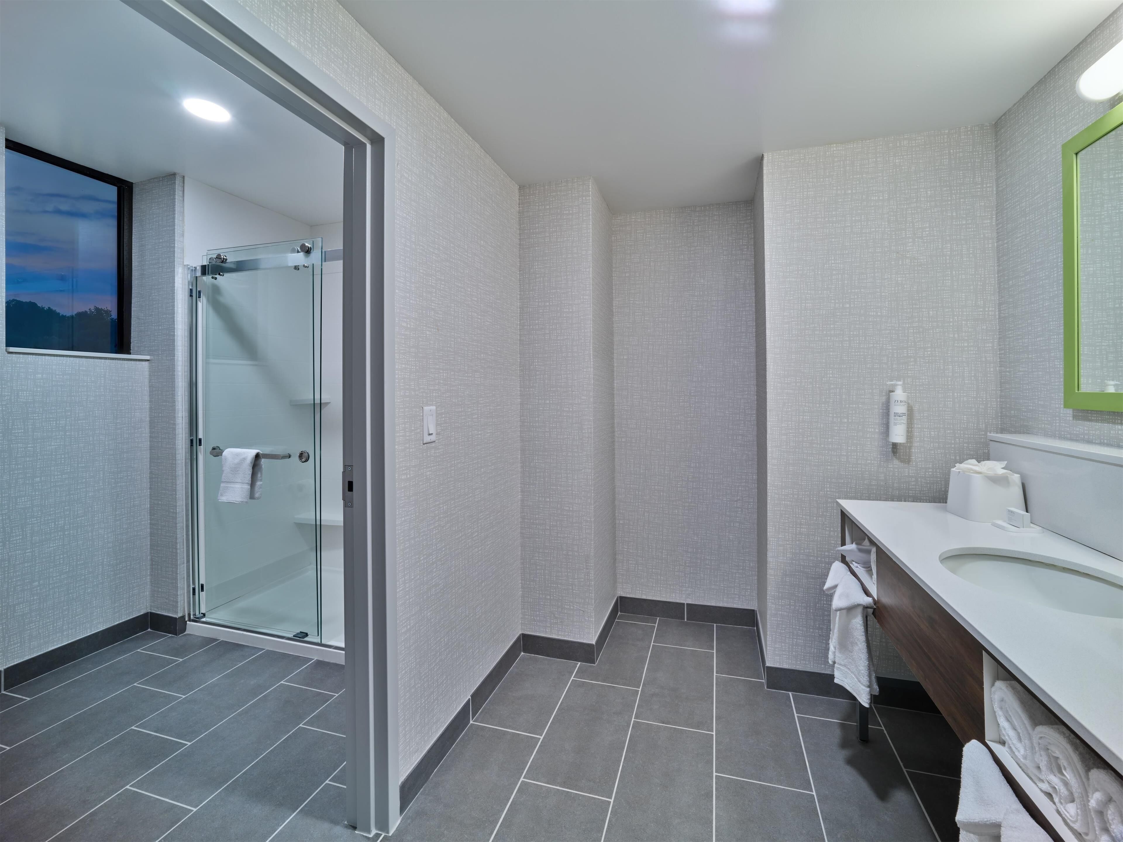 Un baño moderno con cabina de ducha de vidrio, un lavabo blanco con espejo y suelo de baldosas grises.