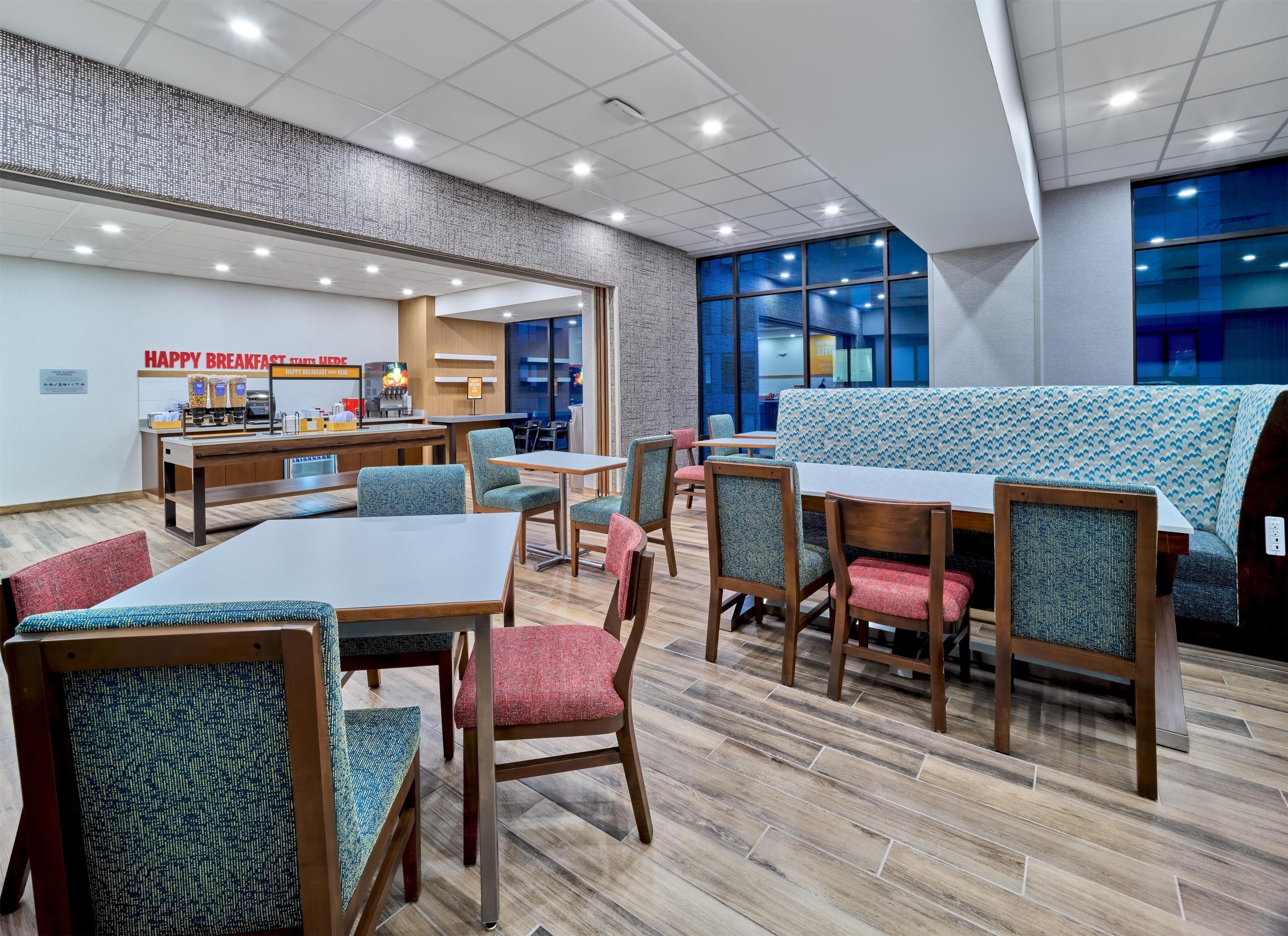 A modern dining area with wooden tables and chairs, a breakfast station, and large windows providing views of the outside.