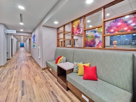 Un pasillo de hotel con piso de madera, un sofá verde con cojines de colores y un cartel que indica los baños.